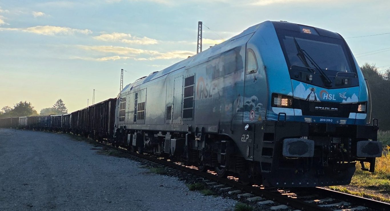 Moderne Lokomotive fährt auf Schienen, umgeben von mehreren Güterwagen im Morgenlicht.